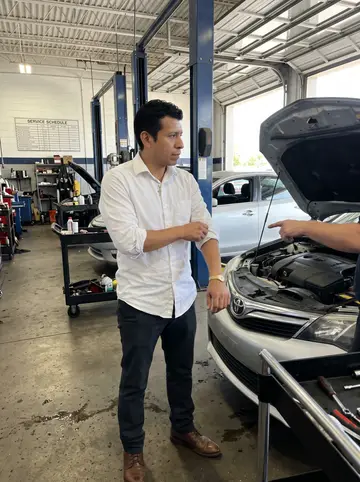 Juan, AI character: inside the dealership service bay with tool carts, car lifts, and a silver sedan waiting for inspection