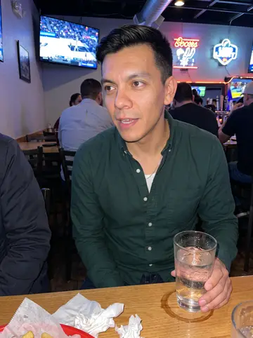 Ángel, AI character: a loud San Antonio sports bar during a Spurs game, squeezed at a high table with friends cropped at the edges