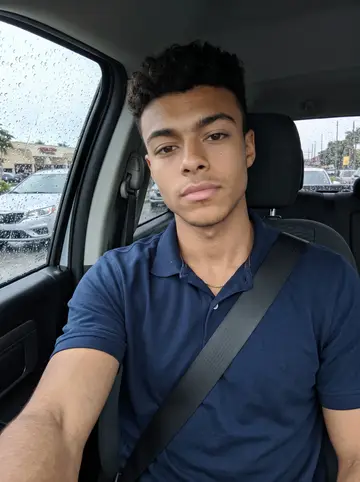 Caio, AI character: inside his parked pickup after a rainy morning route, windshield dotted with water, Hialeah traffic…