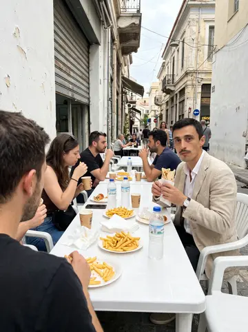 Kostas, AI character: an outdoor table at a taverna in Kastella at night, plates of grilled octopus, bread and a carafe of wine,…