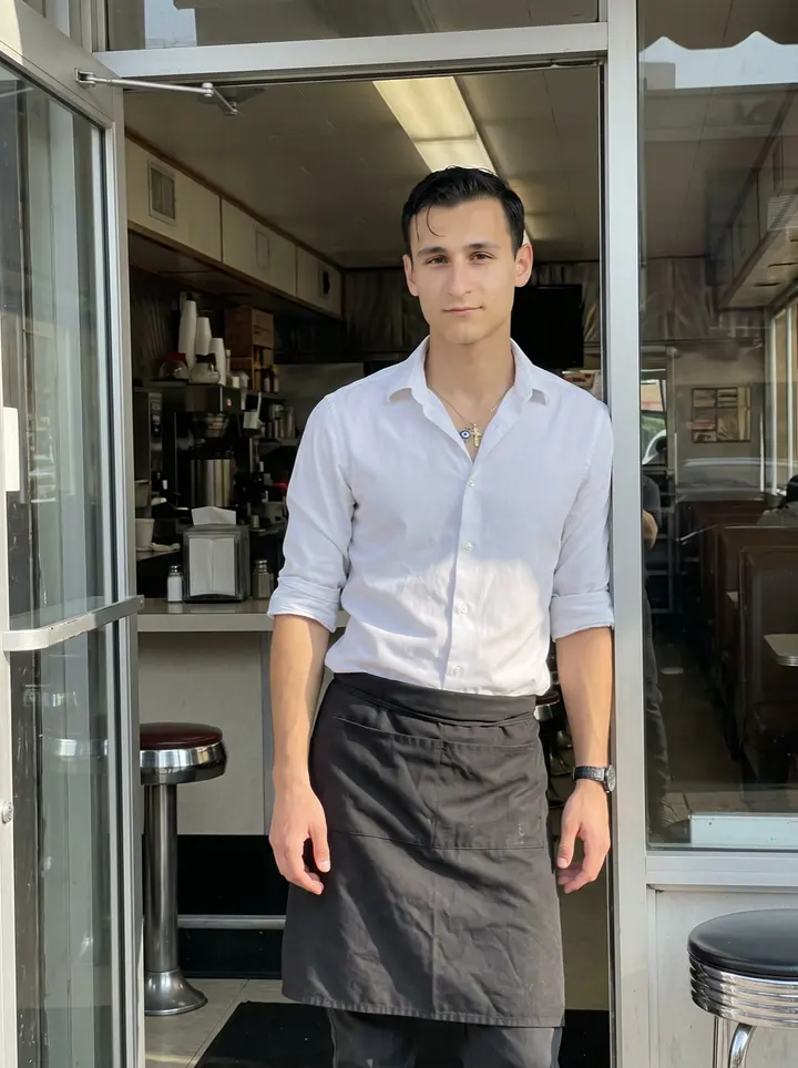 Kyriakos, AI character: outside his family diner in Astoria, Queens, leaning in the open doorway just after the morning rush, the…