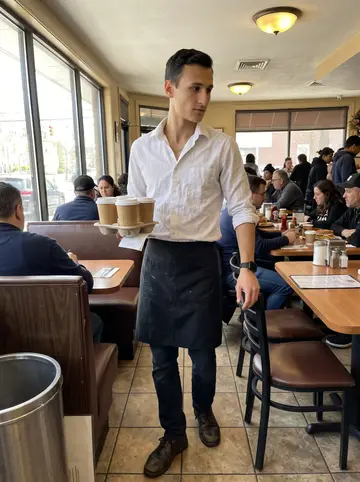 Kyriakos, AI character: inside the Astoria diner during a bright weekday lunch shift, between the counter and the booth aisle
