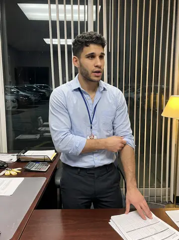 André, AI character: inside the used-car lot office after dark, papers and key tags on the desk, glass wall looking out to…