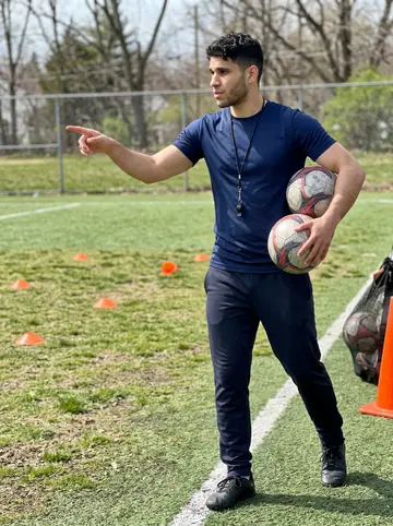 André, AI character: a small Newark soccer field on Saturday morning after youth practice, orange cones and a ball bag on the…