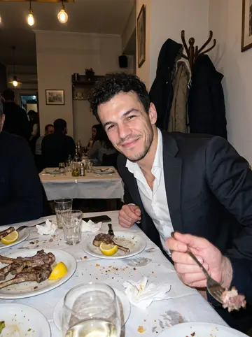 Charalampos, AI character: crowded family baptism dinner in a small Athens restaurant, white tablecloths, plates of lamb and glasses…