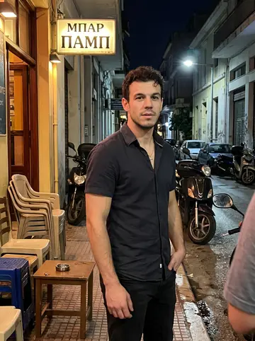 Charalampos, AI character: outside a narrow Athens bar after a humid summer night football match, plastic chairs, low tables and…