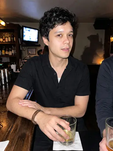 Nikitas, AI character: a crowded Astoria bar after closing shift, with a small TV glowing over the shelves and friends cropped…
