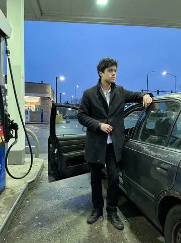 Nikitas, AI character: a Queens gas station beside his uncle's old dark sedan during a late errand, cold pavement shining under…