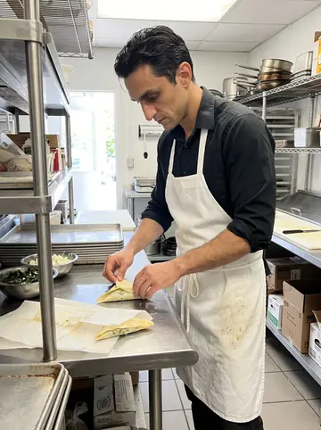 Grigoris, AI character: the restaurant kitchen during prep, stainless counter, trays of spinach and feta, open back door letting…