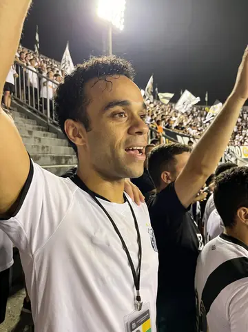 Sol, AI character: the cheap terraces at Vila Belmiro stadium during a Santos FC night match, a crowd of fans blurred behind him