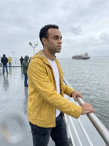 Sol, AI character: the open deck of the Santos–Guarujá ferry in light rain, grey water and a cargo ship in the distance