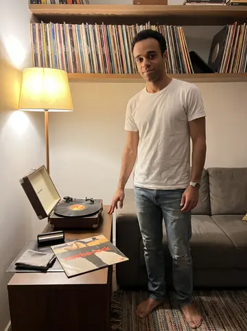 Sol, AI character: the living room of his small apartment in Santos, a shelf full of vinyl records and a battered portable…