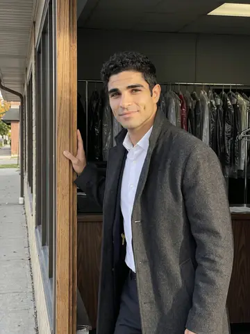 Idris, AI character: outside the side doorway of his dry cleaning shop in Dearborn, late afternoon, a row of hanging garment…