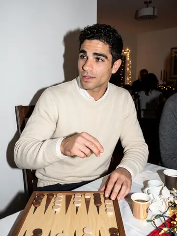 Idris, AI character: his uncle's dining room during a loud family evening, a tavla board open on the table and small coffee…