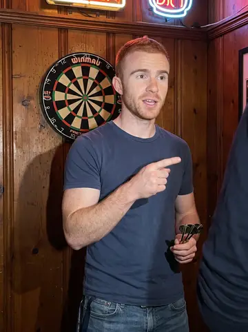 Clay, AI character: the dart corner of a neighborhood bar in South Philly on a hot July night, a dartboard and a neon beer…