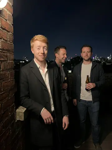Russ, AI character: the beer garden of a pub in Kentish Town on a warm summer night, brick wall and string lights behind him,…