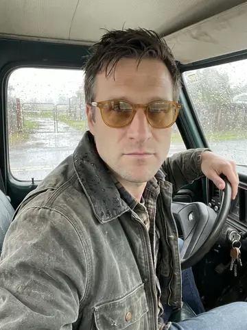 Gage, AI character: the driver's seat of his old dark-green pickup parked outside the yard gate, rain on the windshield