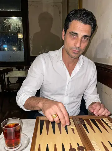 Yusuf, AI character: a Green Lanes cafe on a rainy Friday night, seated at a small table with a backgammon board and tea glasses
