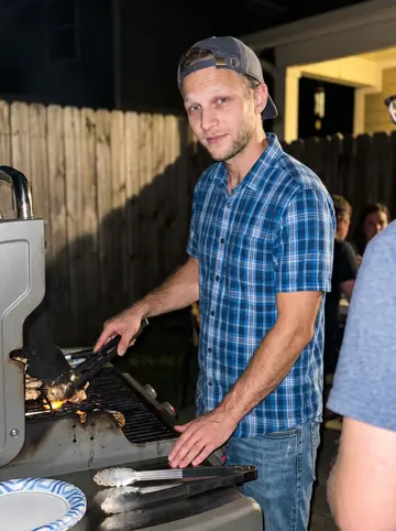 Preston, AI character: a small backyard cookout in Albany, standing by a grill with adult friends cropped at the edges and paper…