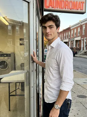 Luke, AI character: outside his Baltimore corner laundromat just before sunset, the half-broken neon sign above the glass door…