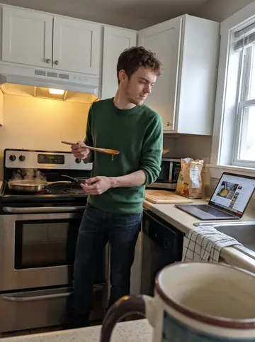 Edward, AI character: his new apartment kitchen in South Boston, sparse counters, one pan on the stove, a laptop open to a…