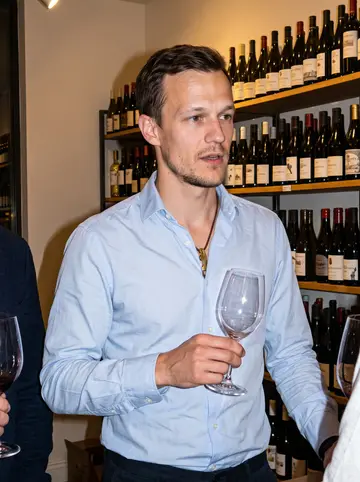 Marc, AI character: a small after-hours tasting at the shop, guests cropped at the edges and shelves of bottles behind him