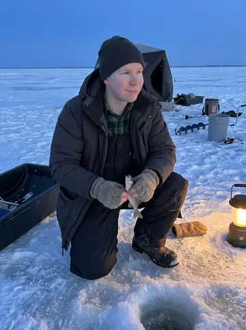 Petro, AI character: on Lake Winnipeg beside an ice fishing hole, a portable shelter and tackle sled behind him, frozen lake…