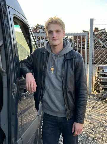 Frederik, AI character: outside the family scrap yard gate in Valby, Copenhagen, leaning against the open door of an old dark van…
