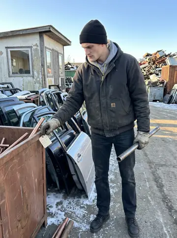 Frederik, AI character: inside the Copenhagen scrap yard between stacked car doors, copper pipe bins and a small weighbridge hut