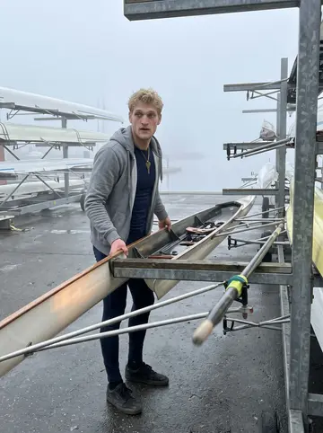 Frederik, AI character: Copenhagen harbour at dawn beside a single scull, low fog over the water and boat racks behind him