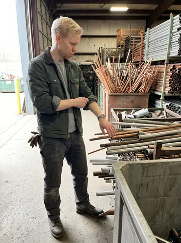 Petri, AI character: inside the recycling yard sorting bay on a clear spring morning, bins of copper pipe and aluminum stacked…