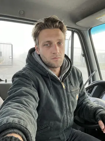 Nils, AI character: parked in the cab of the yard truck before opening, fogged industrial road visible through the side window