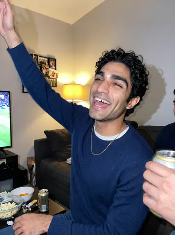 Samir, AI character: his cousin's living room in Dearborn during a Pistons game night, a couch with cushions, snack bowls and…