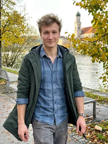 Taras, AI character: the Danube embankment path in Passau in late afternoon, the river and an old-town church tower behind him,…
