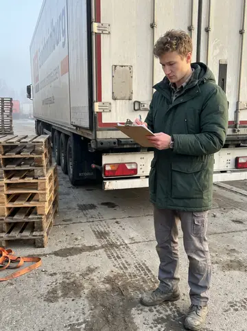 Taras, AI character: the loading dock of a truck yard on the Danube in Passau, wooden pallets stacked behind him, a truck…