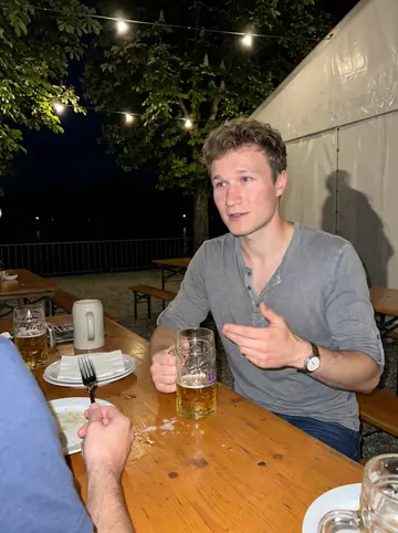 Taras, AI character: a riverside beer garden on the Danube in Passau at night, long wooden tables, steins and plates, string…
