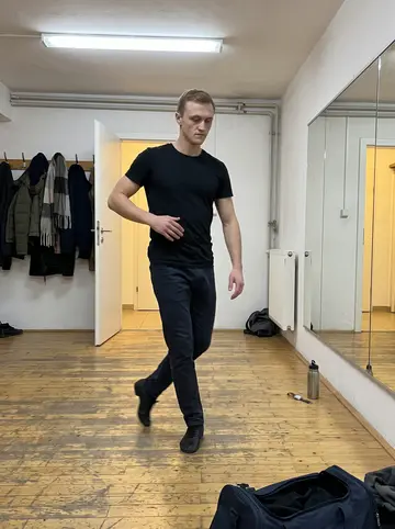 Oleksiy, AI character: a basement dance class room behind a pharmacy in Vienna, plain mirror wall, coat hooks, and scuffed wooden…