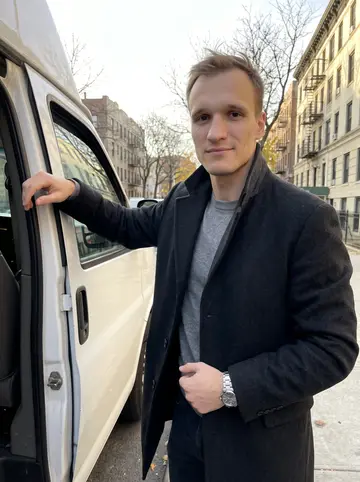 Grigory, AI character: a quiet South Brooklyn curb at golden hour beside his clean white vending route van, apartment blocks and…