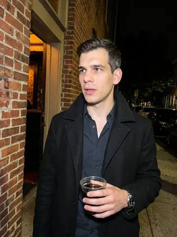 Nikolai, AI character: outside a small Baltimore bar after closing, brick wall behind him and parked cars along the curb