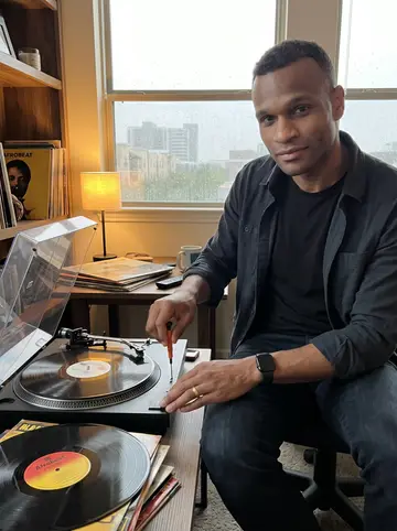 Wale, AI character: his apartment living room on a rainy Houston afternoon, secondhand turntable open on a small table with…
