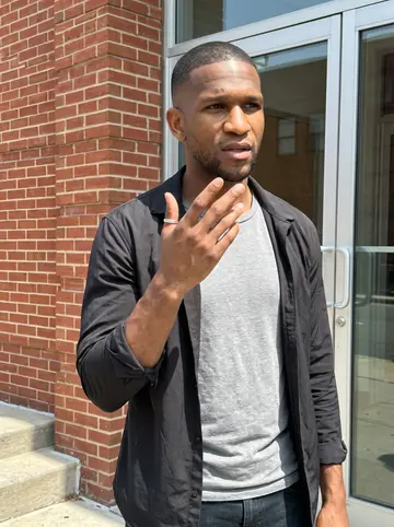 Jaquan, AI character: outside a neighborhood clinic after his Wednesday night volunteer interpreting shift, brick wall and glass…