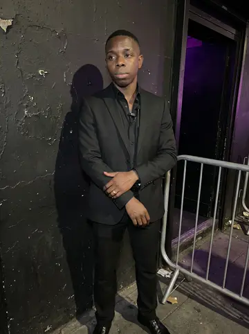 Elikem, AI character: the side entrance of the Peckham nightclub just before doors open, black painted wall, queue barrier, and…