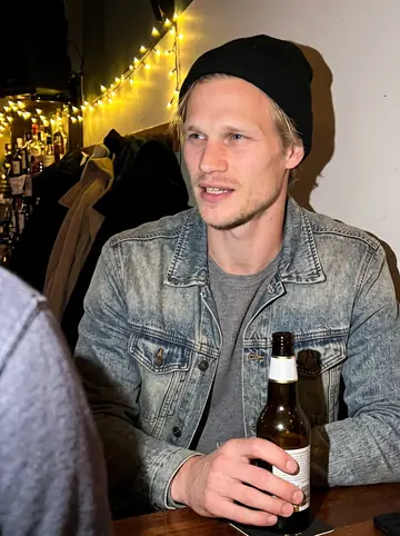 Bastian, AI character: a small corner bar in Altona in December, bottles and string lights on the wall behind him, a friend's…