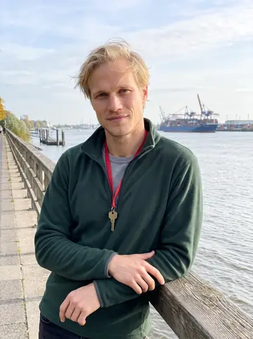 Bastian, AI character: the Elbe promenade near Övelgönne in Hamburg late afternoon, leaning on a weathered wooden railing,…
