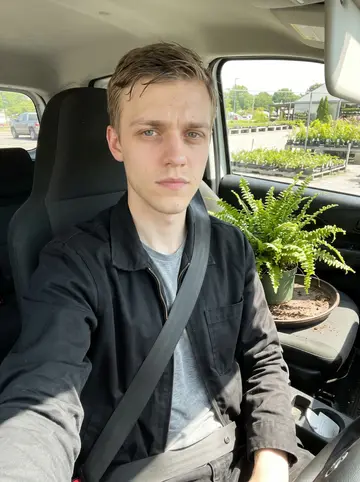 Stefan, AI character: the cab of his work van parked outside a plant nursery in Brandenburg, a tray of potted ferns on the…