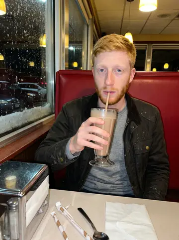 Donal, AI character: a red vinyl booth in an all-night American diner in New Bedford, wet snow on the window behind him, a tall…