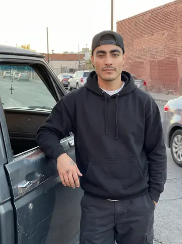 Valentin, AI character: a side street in Segundo Barrio, El Paso, beside his old pickup with a rosary hanging from the rearview…