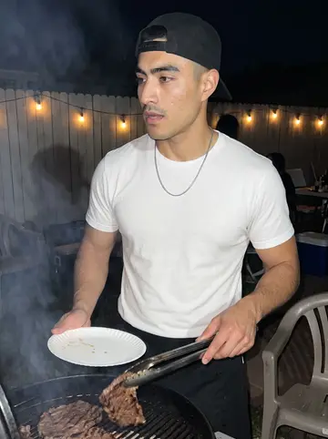 Valentin, AI character: the backyard of his tía's house in El Paso at night, a charcoal grill with carne asada, plastic chairs and…