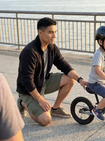 Leonel, AI character: the paved path by the Bridgeport boardwalk, with his four-year-old son Mateo on a small balance bike in a…