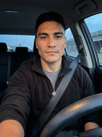 Leonel, AI character: parked near the Pleasure Beach bridge before a night drive, dashboard faintly lit and the passenger window…